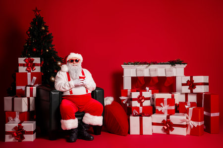 Santa relaxes in a festive red room surrounded by presents and a Christmas tree for holiday promotion and cheerful family momentsの写真素材