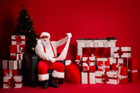 Santa reads a long list among gifts beside a Christmas tree against a bold red backdrop with wrapped presents and festive decorの写真素材
