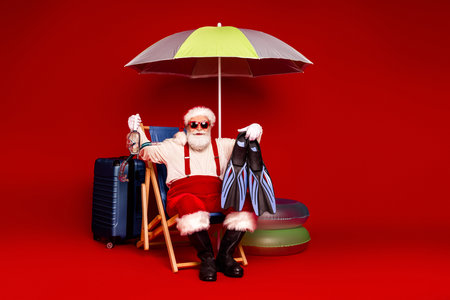 Santa Claus relaxes in a bright red studio with luggage and an umbrella preparing for Christmas season travel and festive shopping fun for family, friends, and home loversの写真素材