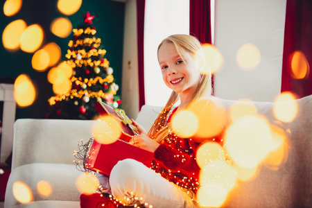 Funny Christmas moment as a girl sits on a sofa opening gifts beside a twinkling tree in a cozy living room full of lights and joyの写真素材
