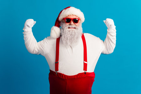 Santa in red suit with suspenders and hat poses proudly against blue background wearing stylish red sunglasses for a festive holiday themeの写真素材