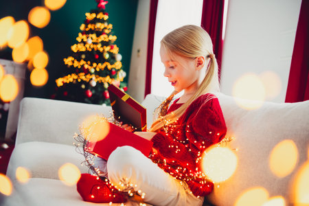 Bright christmas magic at home a girl in a red sweater opens a gift beside the twinkling tree and warm lightsの写真素材
