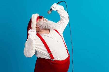 Christmas performance by Santa in red suspenders singing into a microphone against a blue backdrop for festive campaignsの写真素材