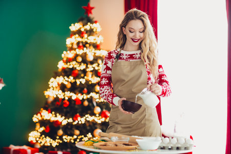 Funny holiday baker in cozy home pours milk for cookies beside a magical Christmas tree and sparkling lightsの写真素材