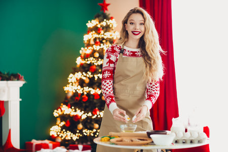 Funny festive kitchen scene cheerful blonde woman bakes cookies beside a twinkling Christmas tree during a cozy home celebrationの写真素材