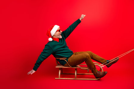 Cheerful Santa on a retro sled joins a merry holiday moment wearing a green sweater and red hat against a bold red backgroundの写真素材