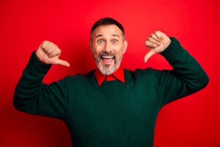 Funny cheerful mature man in green sweater thumbs up and thumbs down against red background perfect for christmas and holiday seasonの写真素材
