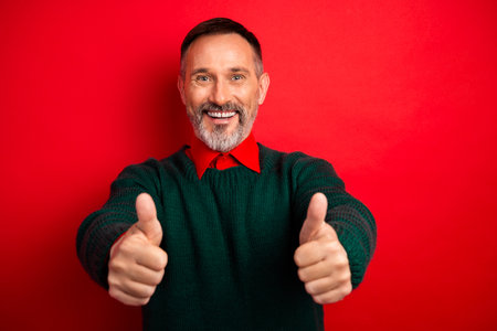 Funny cheerful man in green sweater thumbs up against bold red background perfect for holiday season promotions and Christmas inspired campaigns involving joy celebration and friendshipの写真素材