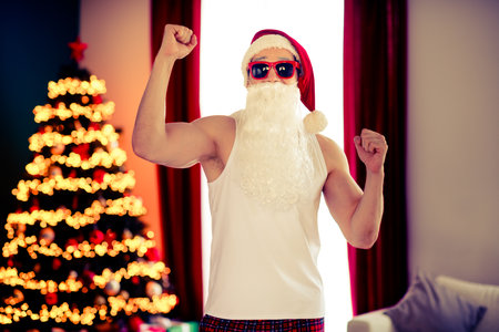 Festive Santa in casual attire posing confidently by the Christmas tree inside a warmly lit holiday-decorated living room.の写真素材