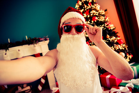 Funny Santa Claus Taking a Selfie With Sunglass by the Festive Christmas Tree and Decorated Living Roomの写真素材