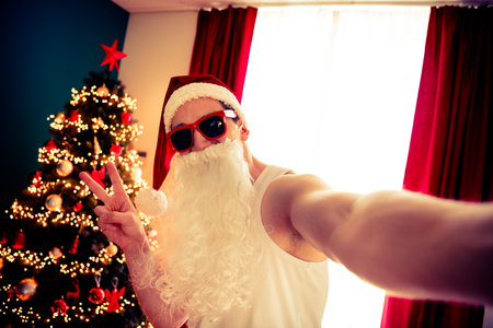 Funny Santa Taking a Festive Selfie With Sunglass and Beard by Christmas Tree in Cozy Home Interiorの写真素材