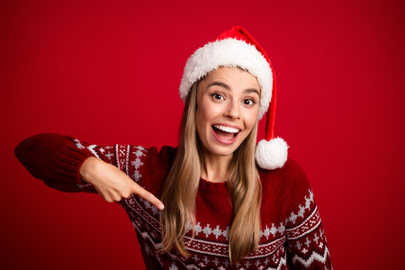 Playful young woman in a Santa hat and red sweater points with a big smile against a bold red background for festive holiday stock photographyの写真素材