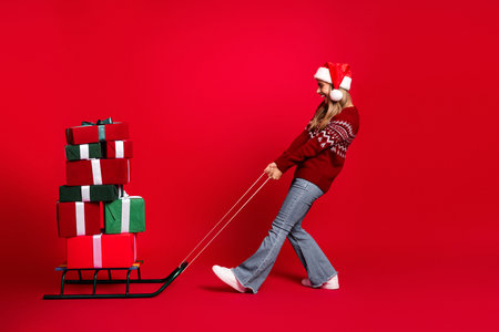 Cheerful woman in a santahat pulls a sled stacked with gifts in a bright red festive scene with holiday joy and Christmas spiritの写真素材