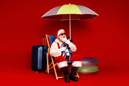 Santa Claus in sunglasses relaxes on a beach chair under a bright umbrella beside luggage and inflatable ringsの写真素材