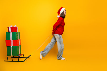 Funny christmas moment girl in red sweater pulls a gift sled with stacked presents across a bright yellow background celebrating x mas seasonの写真素材