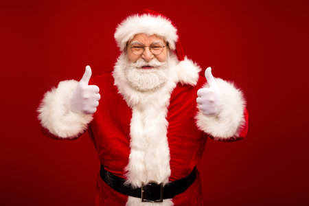 Santa Claus gives thumbs up in red suit with white fur trim smiling for holiday photo against red backgroundの写真素材
