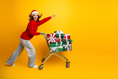 Cheerful santa hat wearing shopper pushes a cart loaded with gifts through a bright yellow studio smiling and laughing ready for Christmas shopping and holiday funの写真素材