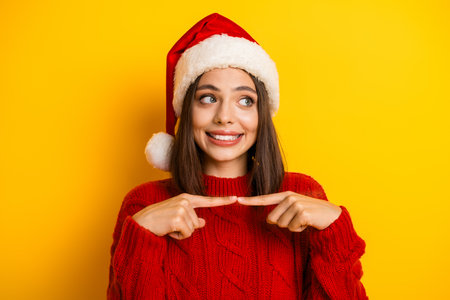 Fun festive moment a pretty young woman in Santa hat points to the space beside her against yellow background while smiling and feeling merry and playfulの写真素材
