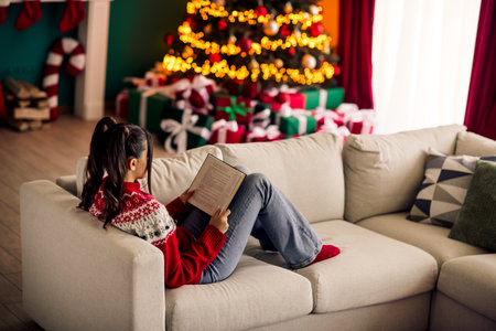 Young woman reading a book on a cozy couch during Christmas holiday season in a festive decorated room with a treeの写真素材