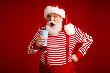 Santa in striped shirt with suspenders drinks from a blue cup smiling in a festive red studio backgroundの写真素材