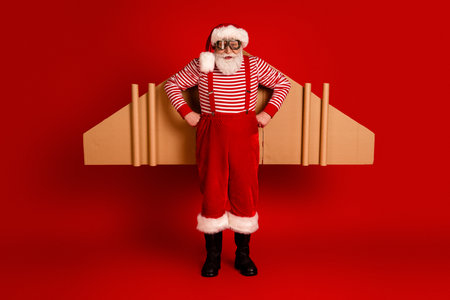Santa in striped shirt red pants suspenders and cardboard wings poses happily against a bold red background for festive stock imageの写真素材