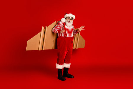 Santa in a striped red and white shirt with suspenders carries cardboard rocket wings on a vivid red studio backdrop for a festive holiday stock photoの写真素材