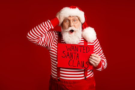 Santa Claus holds a wanted sign in a striped red and white outfit with suspenders against a bold red background for Christmas holidayの写真素材