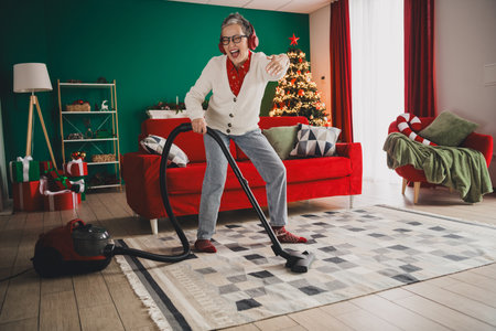 Elderly woman enjoying the holiday spirit, vacuuming her cozy festive home with joy, surrounded by Christmas decorationsの写真素材