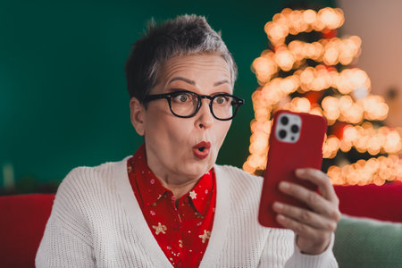 Surprised woman with phone in cozy home during festive Christmas season, showcasing a joyful and celebratory holiday spiritの写真素材