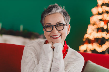 Charming middle-aged woman in cozy indoor setting with festive Christmas tree and decorations, radiating joy and happinessの写真素材