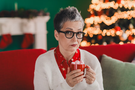 Charming senior woman enjoying Christmas festivities at home surrounded by festive decorations and a holiday treeの写真素材