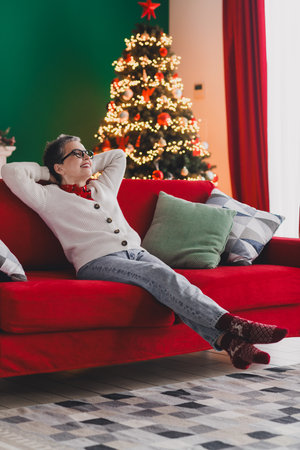Elderly woman relaxing on a red sofa with a cozy Christmas tree backdrop, celebrating the festive holiday spirit in styleの写真素材