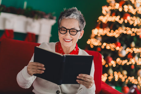 Charming elderly woman reading a book by a Christmas tree in a cozy festive home decorated for the holiday seasonの写真素材