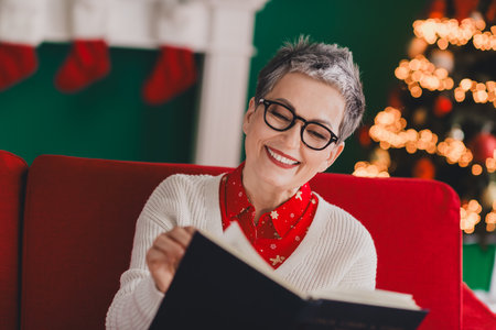 Beautiful older woman reading a book next to a festive Christmas tree in a cozy home during the holiday seasonの写真素材