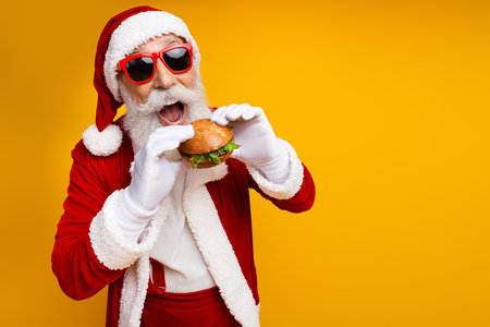 Joyful Santa Claus enjoying a delicious burger in Christmas spirit against a vibrant yellow backgroundの写真素材