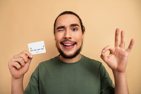 Young man smiling with a credit card and thumbs up gesture in a casual beige studio settingの写真素材