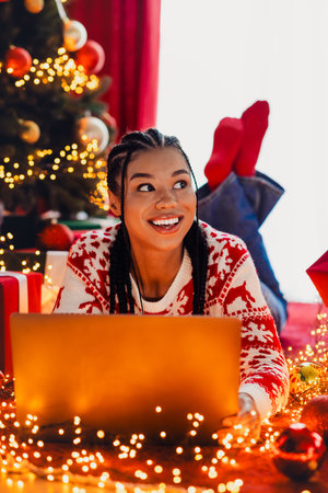 Playful Christmas vibes with a smiling woman on a laptop surrounded by twinkling lights a decorated tree and festive gifts in a cozy home settingの写真素材