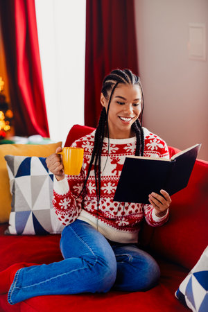 Cheerful woman reads a holiday book on a red couch holding a yellow mug in a cozy Christmas home sceneの写真素材