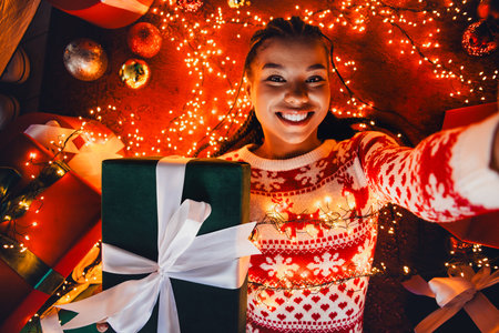 A joyful young woman takes a selfie while holding a large green gift box wrapped in white ribbon amid festive Christmas lights and warm decorations at homeの写真素材