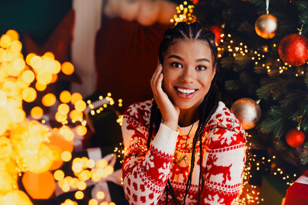 Funny stylish woman with braids by a festive christmas tree smiles in cozy indoor setting with warm lightsの写真素材