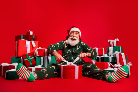 Joyful Santa in festive pajama surrounded by wrapped gifts smiling widely in a bold red studio setting for holiday cheer and Christmas magicの写真素材