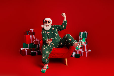 Santa in festive pajama sits in a red chair surrounded by wrapped gifts celebrating Christmas and holiday joy with a smileの写真素材