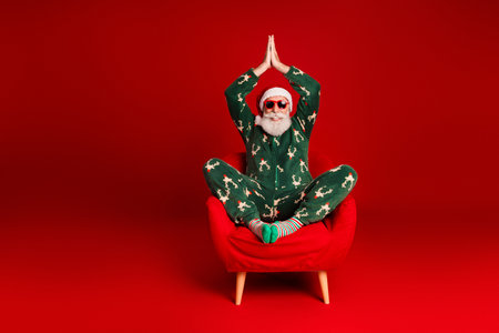 Santa in green Christmas pajama sits relaxingly on a red chair with hands together in a prayer pose against a bold red backgroundの写真素材