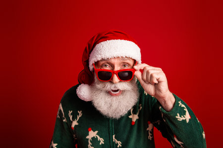 Santa in a festive green reindeer sweater wears red sunglass against a bright red backdrop delivering Christmas cheerの写真素材