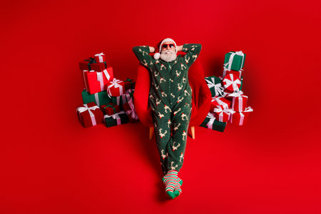 Santa relaxes in festive pajama suit surrounded by gifts against a bold red background for a joyful Christmas sceneの写真素材