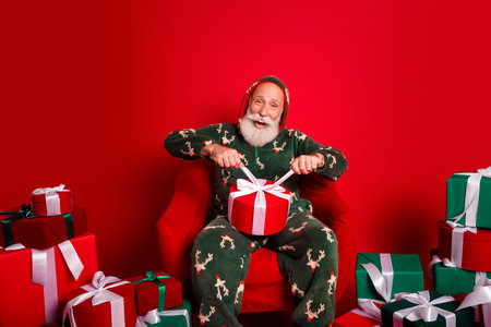 Santa in cozy pajama opens a bright red gift as Christmas joy fills a festive home surrounded by colorful presents and ribbons in a heartwarming holiday sceneの写真素材