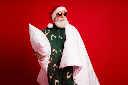 Santa in cozy pajama cape holds pillow smiles under red background for Christmas fun and festive season shopping dreamの写真素材