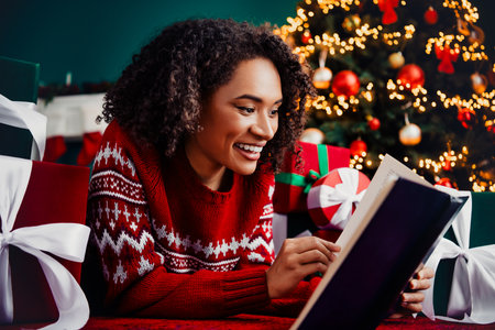 Playful holiday moment as a woman in a red sweater reads a book among gifts and a glowing Christmas tree creating warm home vibesの写真素材