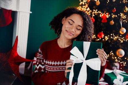 Joyful holiday moment as a woman in a cozy red sweater gives a gift beside a twinkling Christmas tree at home full of warm lightsの写真素材