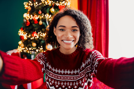 Fun holiday selfie with a smiling woman in a red christmas sweater in a cozy home setting featuring a Christmas tree lightsの写真素材